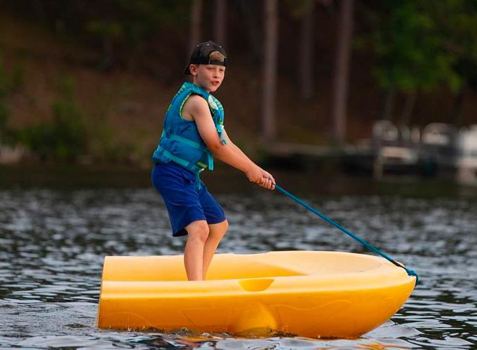 A camper at North Star Camp for Boys, stands on a yellow watercraft, also known as a Fun Bug, while holding onto the pulley in Spider Lake.