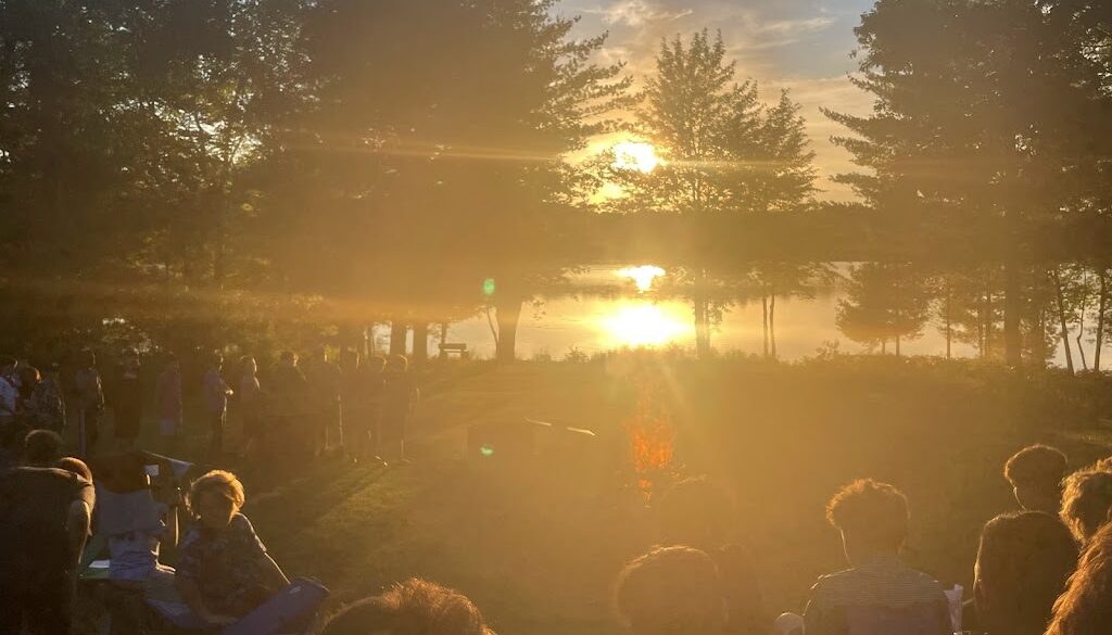 A group of campers and staff members gather around a firepit at sunset to listen to staff members speak at North Star Camp for Boys.