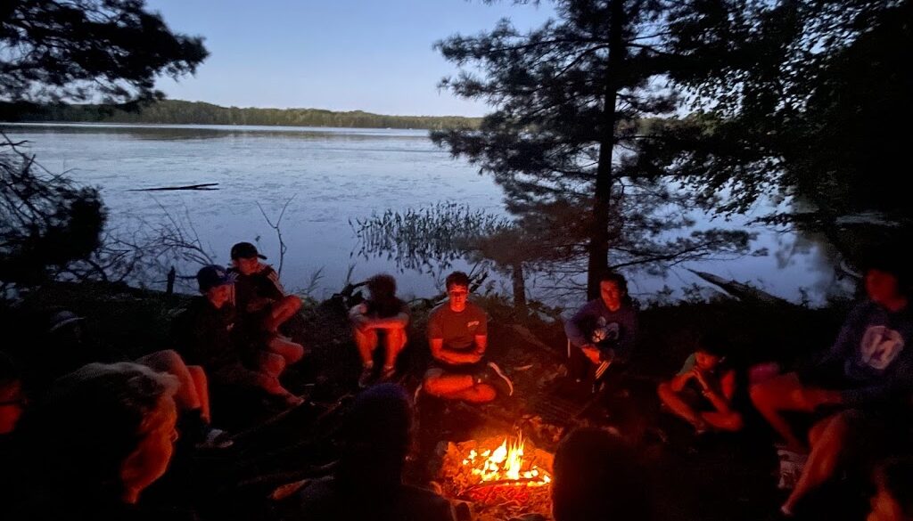 A group of campers and staff members from North Star Camp for Boys sit in a circle around a campfire on the lake shoreline.