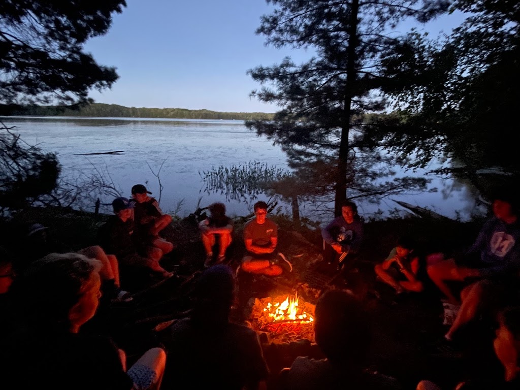 A group of campers and staff members from North Star Camp for Boys sit in a circle around a campfire on the lake shoreline.