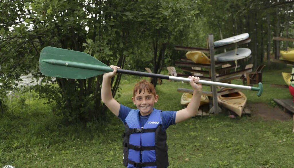 A camper wearing a lifejacket smiles while holding up a canoe ore, canoes are stacked in the background.