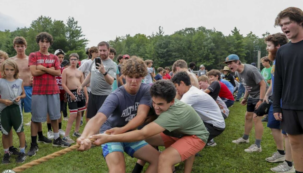 A group of campers participate in an intense game of tug-of-war, while non-participating campers cheer them on.