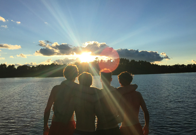 Four campers stand on the lakeshore at North Star Camp for Boys with their arms around each other looking out at Spider Lake as the sun sets.