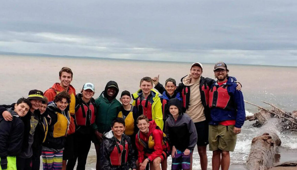 A group of campers and staff members at North Star Camp for Boys smile for a photo on the shoreline during a Wilderness Trip.