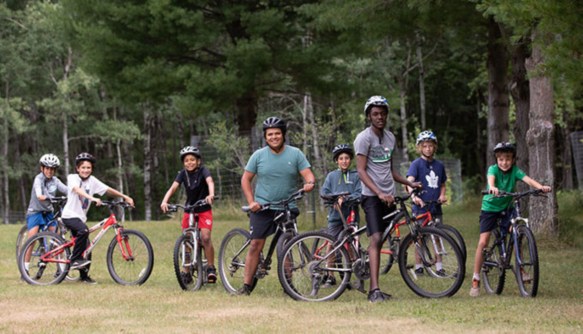 A group of campers and two staff members smile for a photo while enjoying a bike ride through the campgrounds of North Star Camp for Boys.