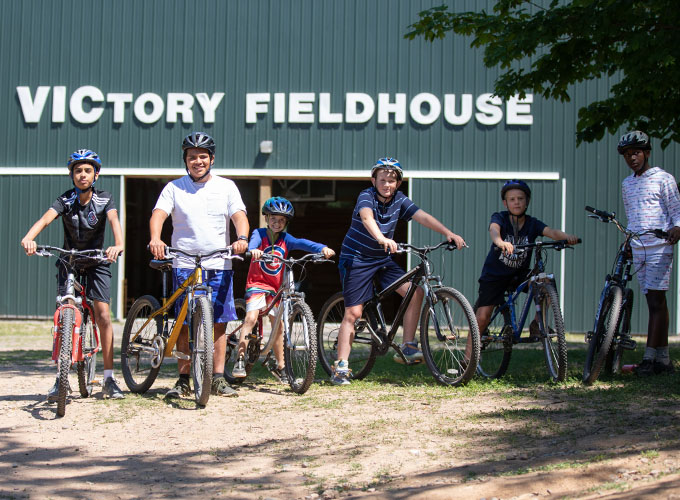A group of five campers and one staff member sit on bikes and wear helmets, while smiling for a picture outside of Victory Fieldhouse at North Star Camp for Boys.