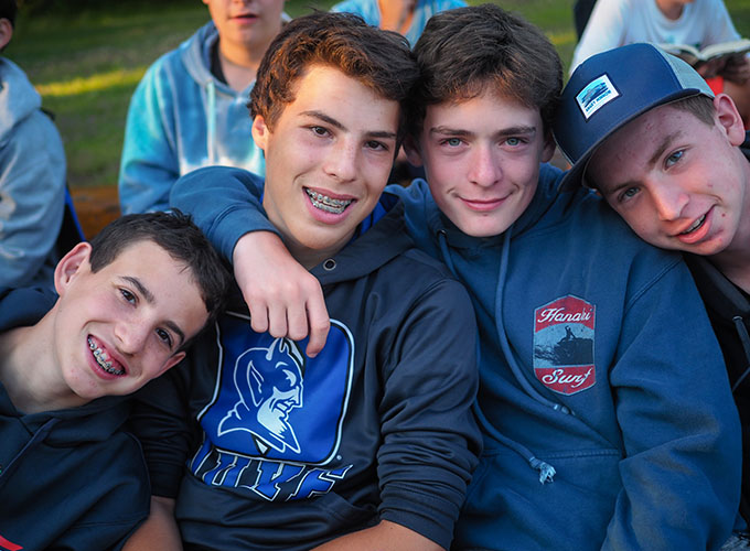 A group of four high school-aged boys smile for a picture at North Star Camp for Boys. One has his arm around the other, and the other two are leaning in.