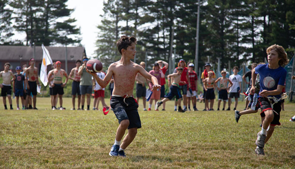 A camper prepares to throw a football, while another camper runs beside him. A group of campers stand behind them watching the game at North Star Camp for Boys.