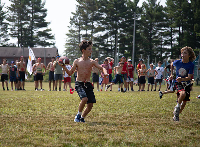 A camper prepares to throw a football, while another camper runs beside him. A group of campers stand behind them watching the game at North Star Camp for Boys.