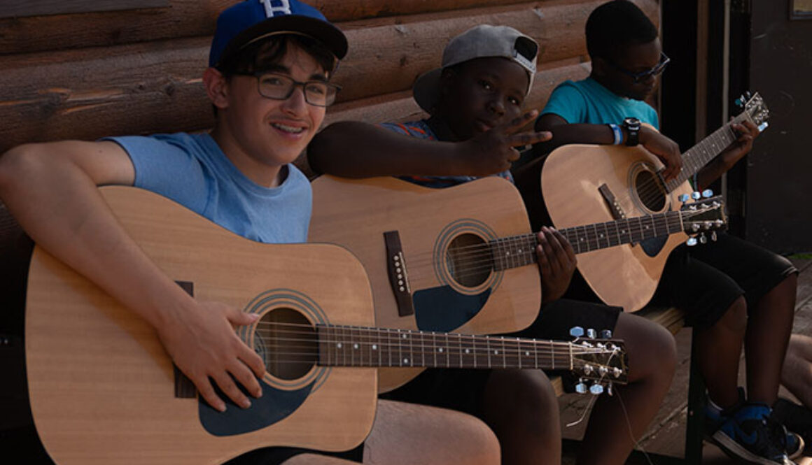 Three campers sit outside of a cabin while strumming guitars at North Star Camp for Boys.