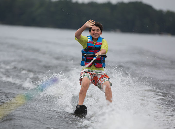 A camper smiles and waves as he holds onto a tow rope while waterskiing through Spider Lake at North Star Camp for Boys.