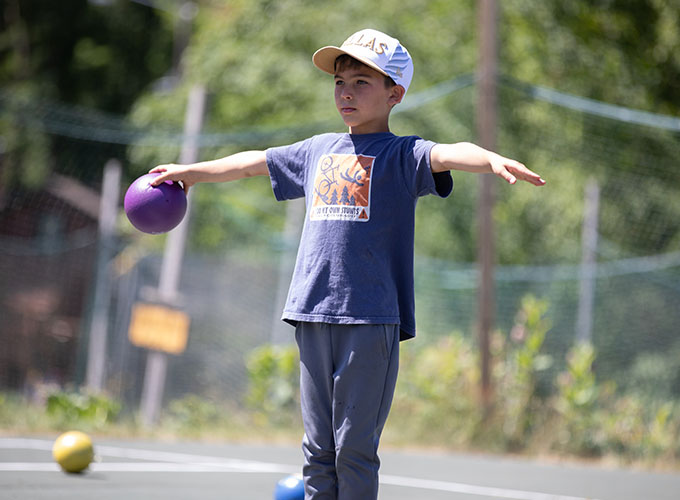 A boy holds out his arms, creating a "T" shape. He is holding a purple foam ball in one hand and looks onward.