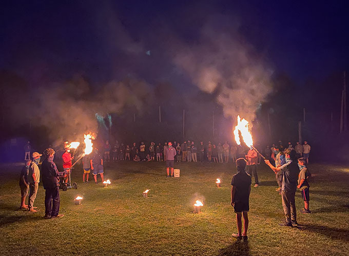 A group of campers stand in a semi-circle while facing each other, two boys on each side hold flaming torches. Another group of counselors and campers watch from the distance.