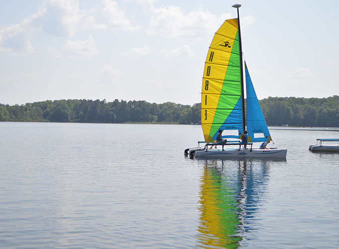 Three campers leisurely cruise on a yellow, green, and blue sailboat in Spider Lake at North Star Camp for Boys.