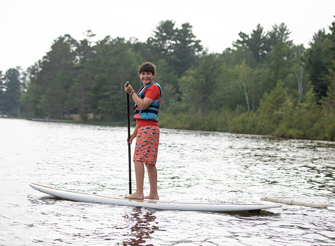 A camper smiles while stand up paddle boarding in Spider Lake at North Star Camp for Boys.