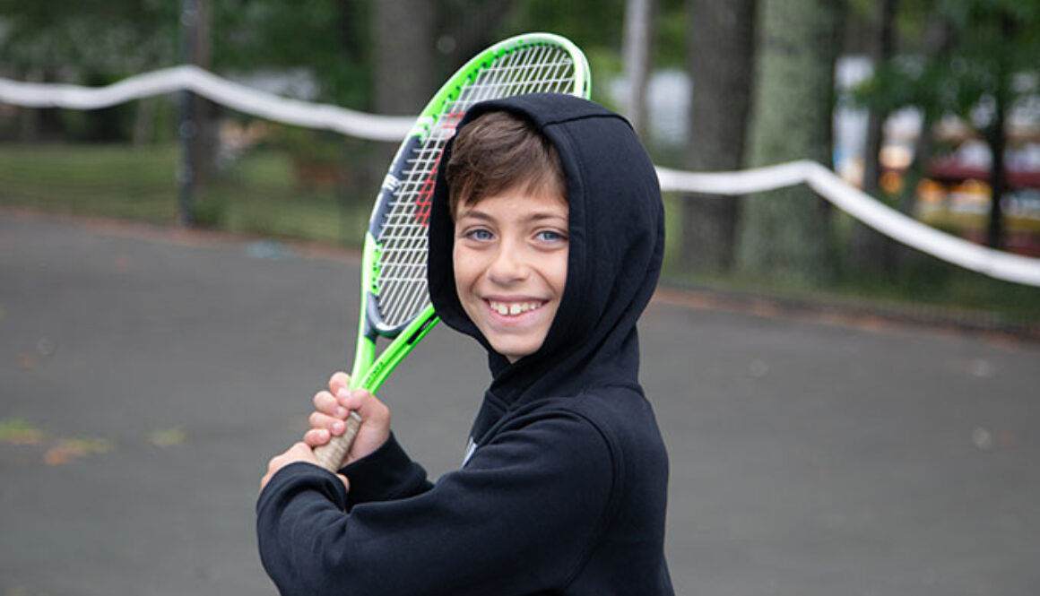 A camper at North Star Camp for Boys smiles and holds up a tennis racquet.