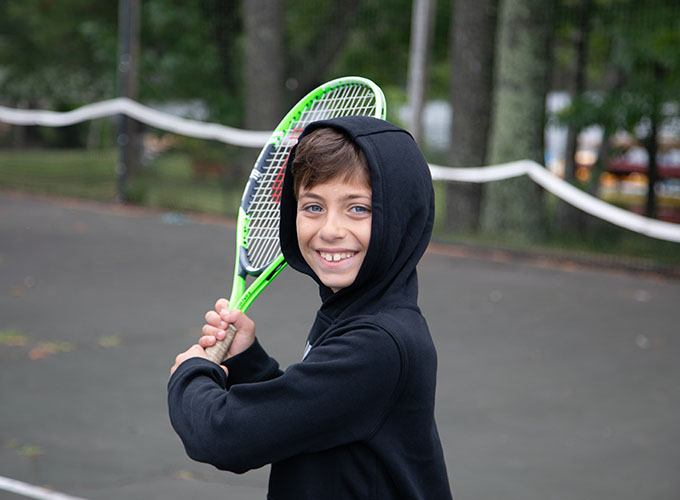 A camper at North Star Camp for Boys smiles and holds up a tennis racquet.
