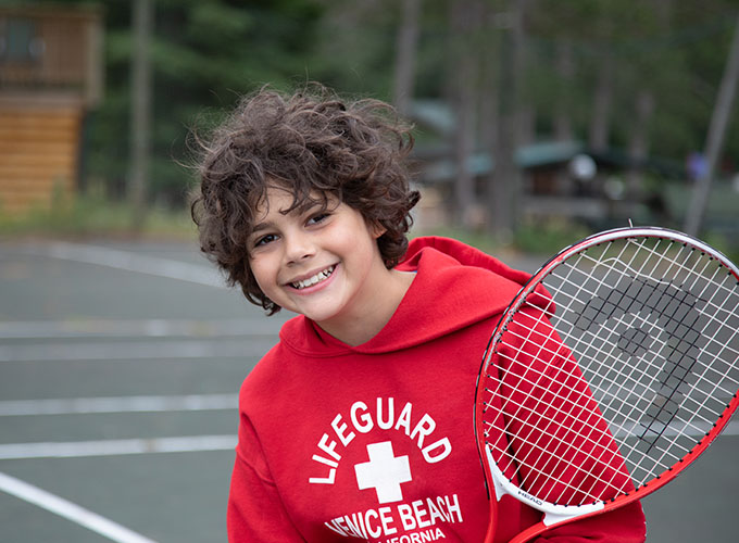 A camper at North Star Camp for Boys smiles while wearing a lifeguard sweatshirt, and holds up a tennis racquet.