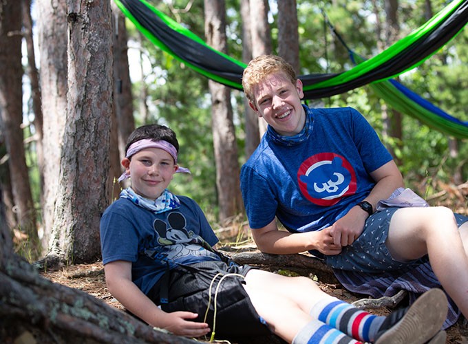 A North Star Camp for Boys camper and staff member smile while sitting in the woods together, hammocks are hanging from trees behind them.