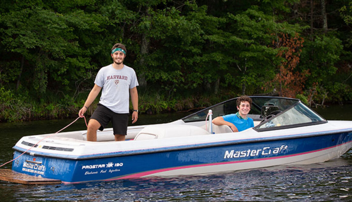 Two North Star Camp for Boys staff members smile while in a boat, one is sitting in the drivers seat, and the other is standing holding the tow rope.