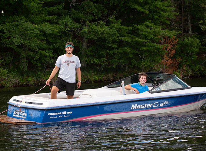 Two North Star Camp for Boys staff members smile while in a boat, one is sitting in the drivers seat, and the other is standing holding the tow rope.