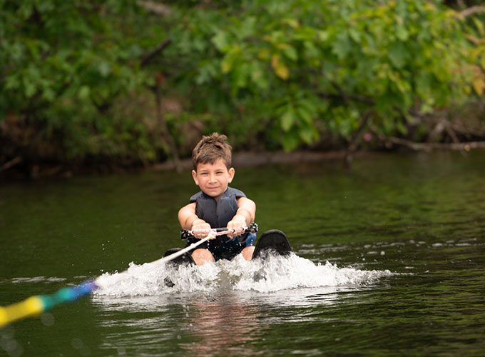 A young camper wearing water skis sits in Spider Lake while holding onto a tow rope, as he is gently pulled through the water during a waterskiing lesson at North Star Camp for Boys.