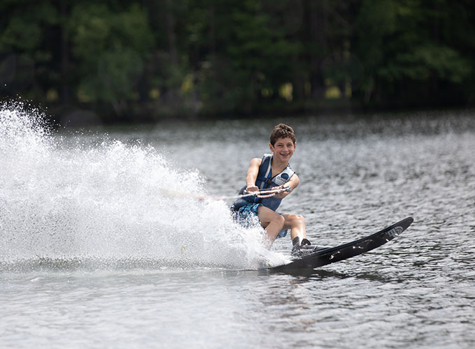 A camper smiles as he navigates slalom waterskiing, leaving a fountain-like burst of water behind him in Spider Lake at North Star Camp for Boys.
