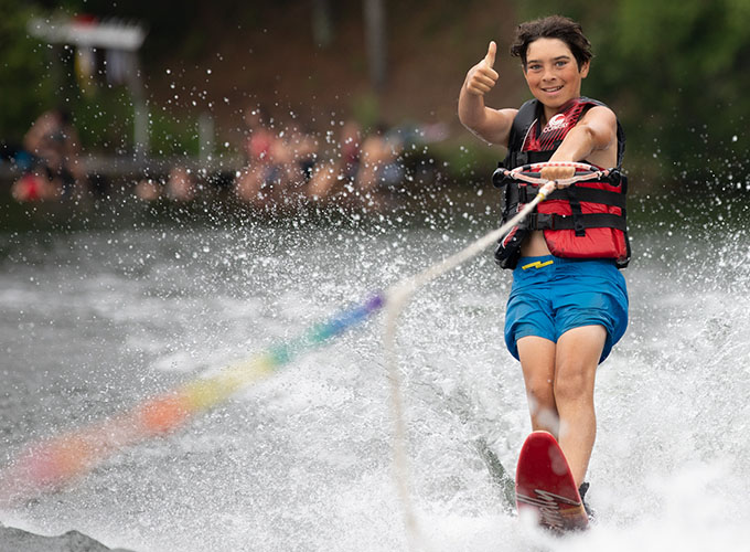 A camper smiles while giving a thumbs up motion as he slalom waterskies through Spider Lake at North Star Camp for Boys.
