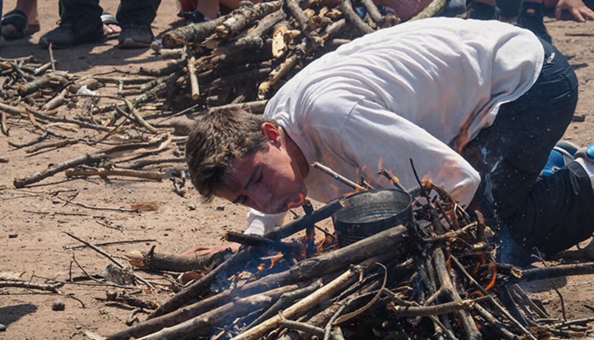 A camper kneels on the ground while blowing on wood in an effort to start a fire during a competition at North Star Camp for Boys.