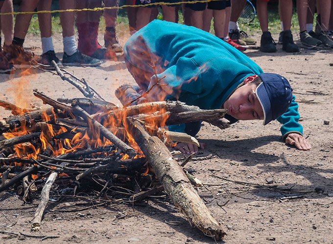 A camper kneels on the ground and blows on wood to enhance the slight fire that is building during a fire starting competition at North Star Camp for Boys.