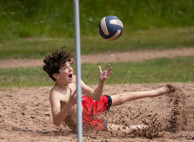A camper with a shocked facial expression dives into the sand as he watches a volleyball fly towards him, his hands ready to catch the ball at North Star Camp for Boys.