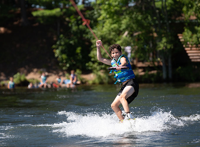 A camper holds a tow rope with one hand as he puts the other in the air while wakeboarding through Spider Lake at North Star Camp for Boys.