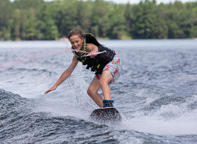 A camper holds a tow rope with one hand, and smiles as he balances on a wakeboard around Spider Lake at North Star Camp for Boys.