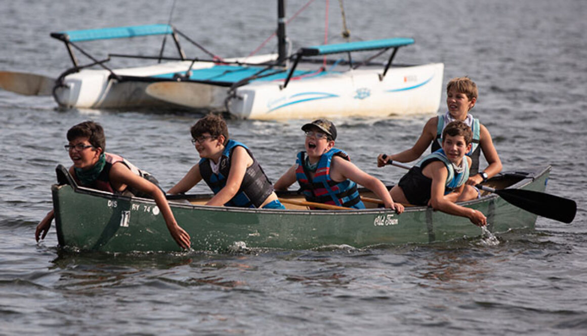 Five North Star Camp for Boys campers all look in the same direction while in a canoe on Spider Lake. The camper in the back holds an ore, while the four other campers have their hands in the water.