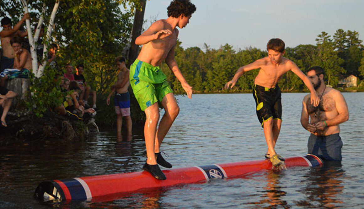 An instructor wades in Spider Lake supervising two campers moving on the water logroll at North Star Camp for Boys.