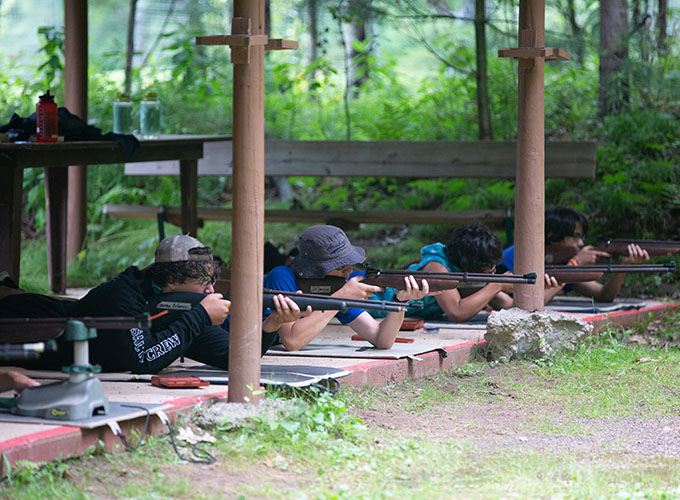 A group of boys aim their rifles while laying on the ground practicing riflery skills at North Star Camp for Boys.