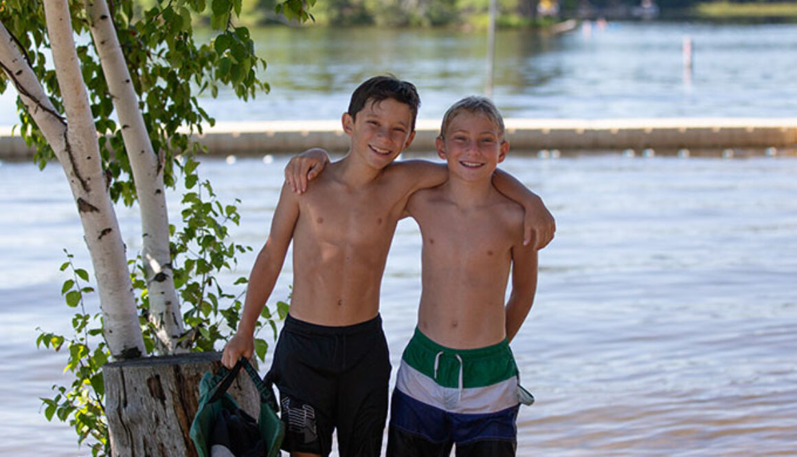 Two campers at North Star Camp for Boys smile with their arms around each other. They are wearing swim trunks and standing in front of Spider Lake.