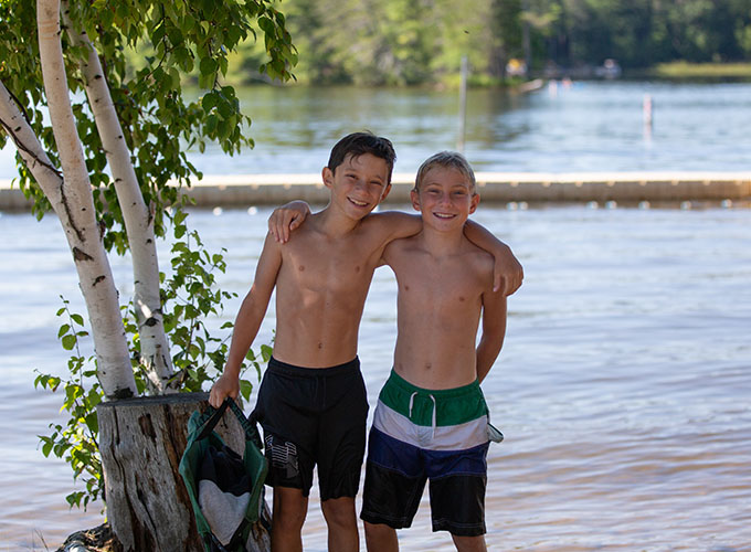 Two campers at North Star Camp for Boys smile with their arms around each other. They are wearing swim trunks and standing in front of Spider Lake.