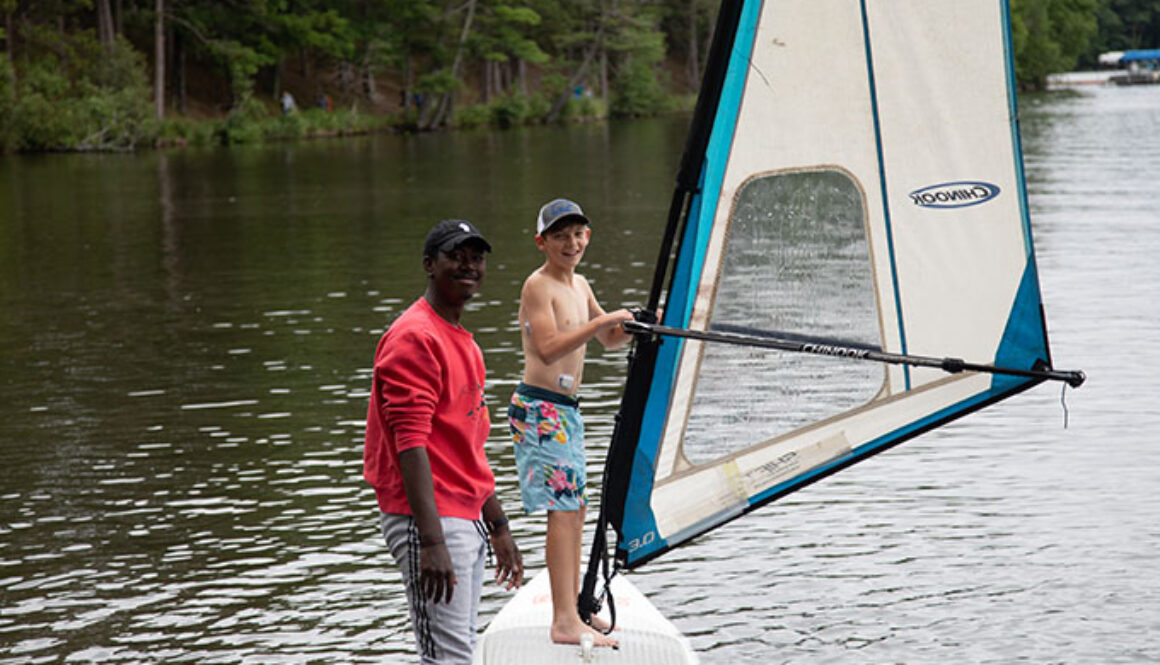 A North Star Camp for Boys camper stands on a windsurf in Spider Lake, while and instructor wades in the water beside him, both are smiling.