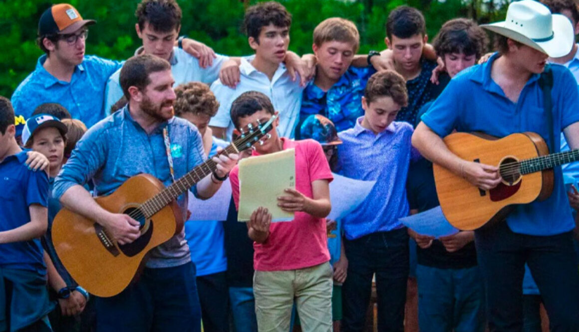 A group of campers and staff members at North Star Camp for Boys sing a song with their arms around each other. Two staff members are playing the guitar.