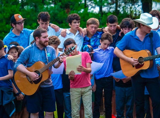 A group of campers and staff members at North Star Camp for Boys sing a song with their arms around each other. Two staff members are playing the guitar.