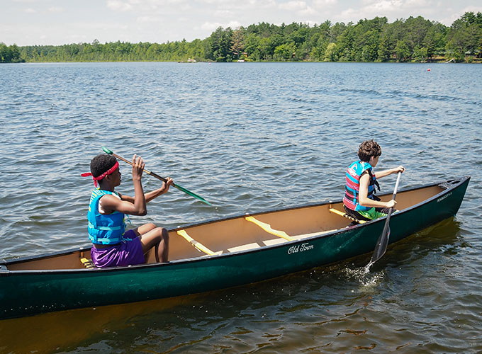 Two campers sit in a tandem canoe and paddle down the river at North Star Camp for Boys.