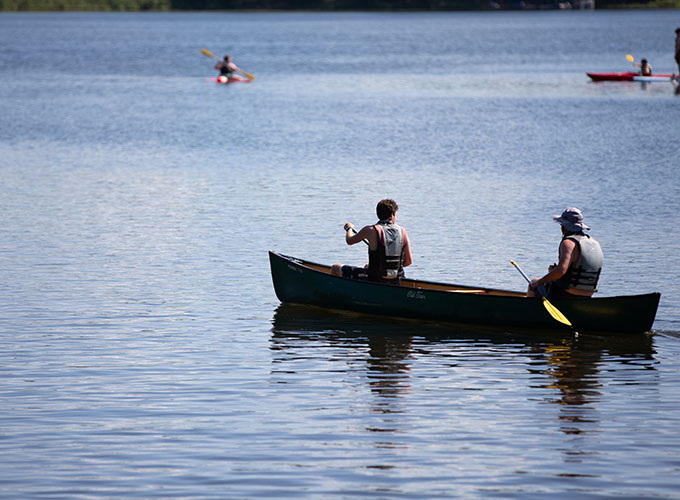 Two staff members wearing life jackets, sit in a tandem canoe and and row through the lake at North Star Camp for Boys.