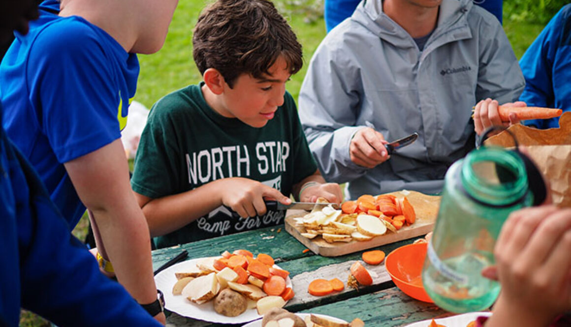 A group of campers peel and slice carrots and potatoes on a picnic table during an outdoor cooking activity at North Star Camp for Boys.