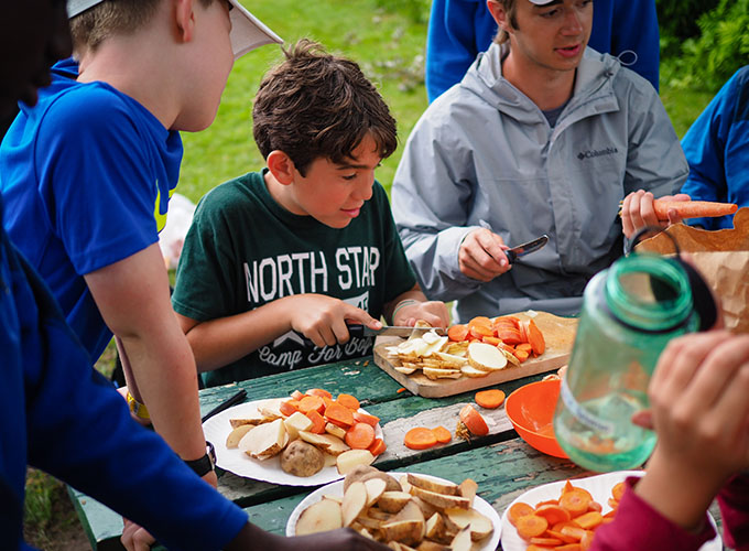 A group of campers peel and slice carrots and potatoes on a picnic table during an outdoor cooking activity at North Star Camp for Boys.