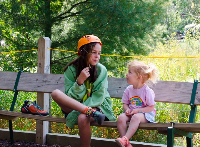 A girl wearing an orange helmet sits on a bench talking to a toddler-aged girl wearing a pink rainbow shirt on the North Star Camp for Boys campground.