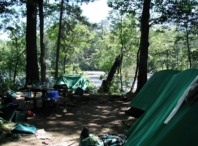 Tents are pitched in the forest, the river can be seen in the distance during a wilderness trip for the campers of North Star Camp for Boys.