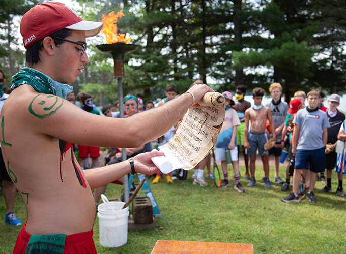 A camper wearing body paint reads from a scroll near a torch flame while campers stand and watch at North Star Camp for Boys.