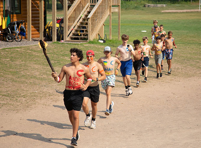 Campers wearing body paint run in a single file line for United Nations Day at North Star Camp for Boys. The leader is carrying a flaming torch.