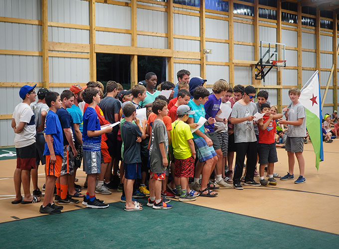 A cluster of campers gather around in Victory Fieldhouse, reading instructions from pieces of paper. One camper is carrying the national flag of Djibouti.